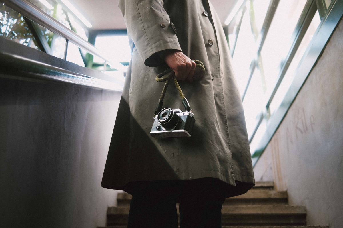 Person in a long coat holding a vintage camera with a rope camera strap on a staircase