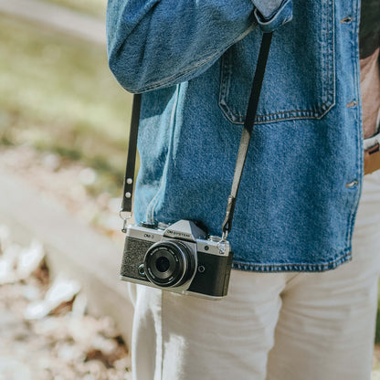 A person standing with the new OM SYSTEM OM-3 camera in a park. The camera has timeless camera strap on it