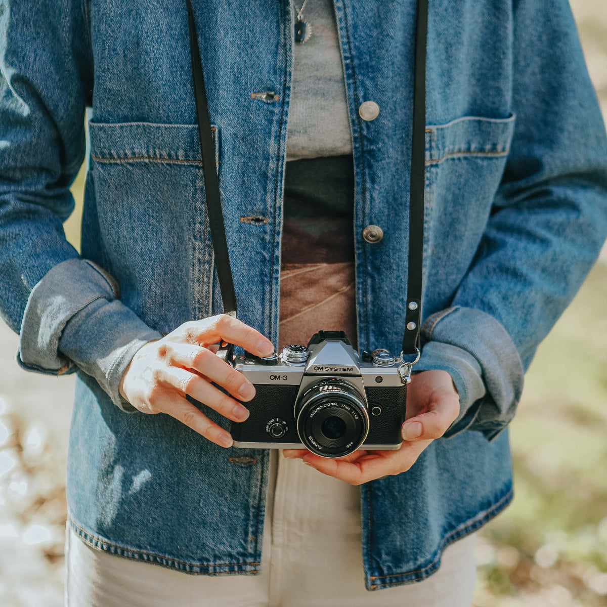 A person outdoors holding the new OM SYSTEM OM-3 camera with black leather camera strap from Arch Appearance