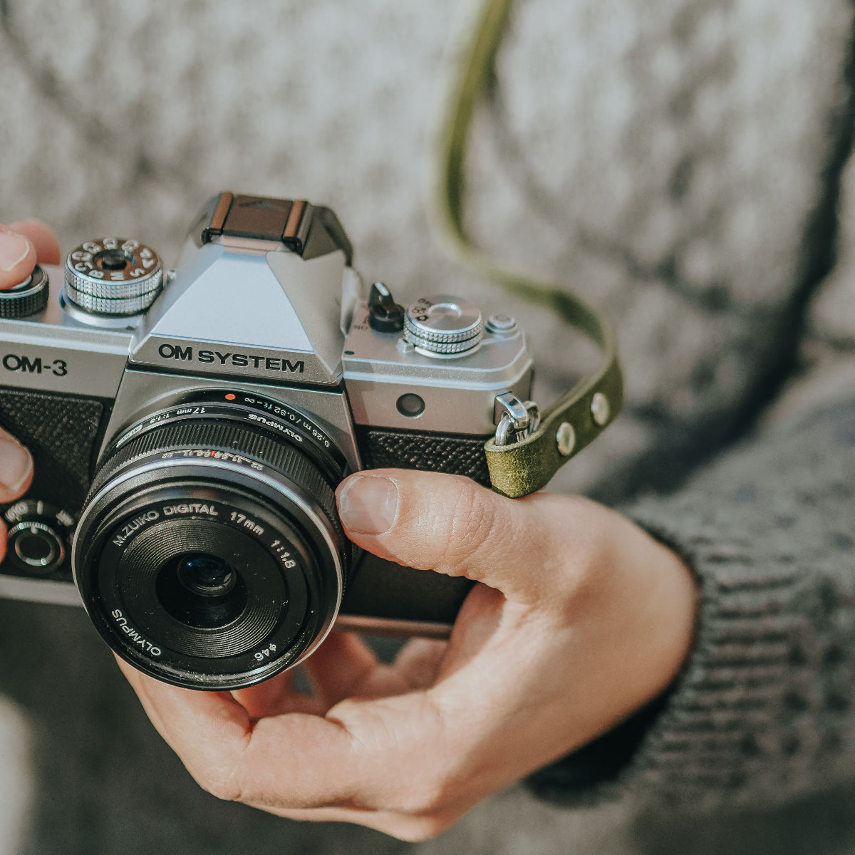 A person holding the new OM SYSTEM OM-3 camera with olive green leather camera strap from Arch Appearance