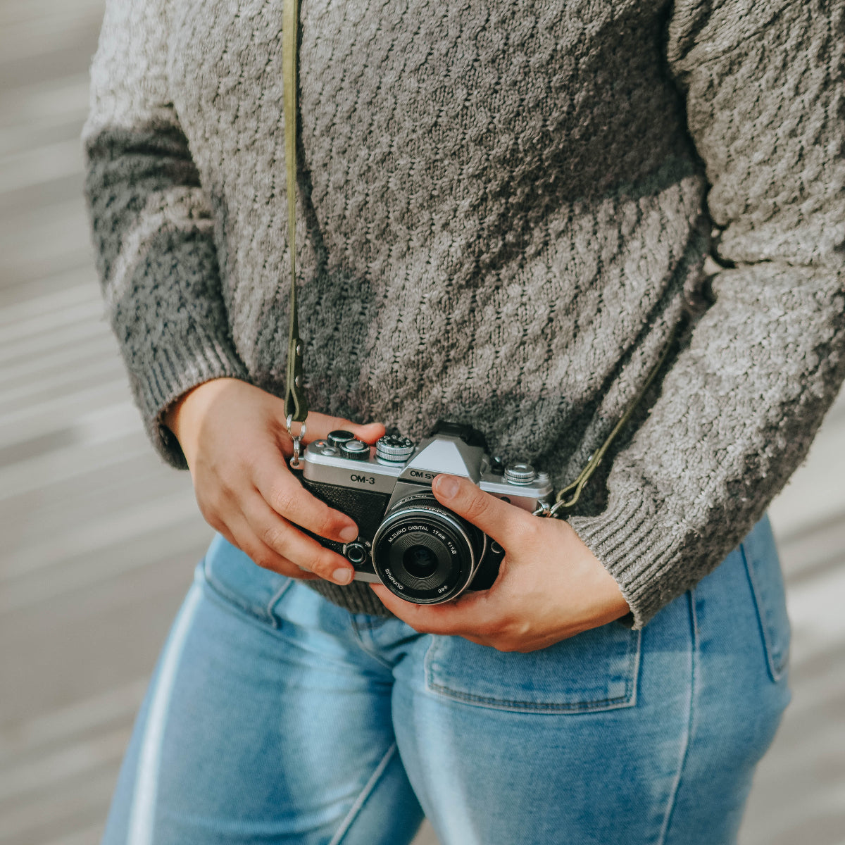 A woman holding the new OM SYSTEM OM-3 camera with olive green leather camera strap which is handmade by Arch Appearance